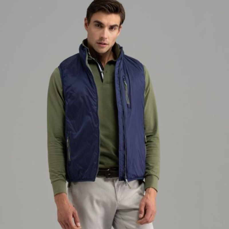 Giovane uomo con polo verde a manica lunga, pantalone chino color grigio ghiaccio con cintura e giubbotto smanicato blu con inserti verdi